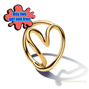 Può includere: Un anello color oro con un design a forma di cuore. L'anello ha una finitura liscia e lucida e uno stile moderno e minimalista. L'anello è mostrato su sfondo bianco. Una bolla grafica rossa e blu con il testo "Buy two get one free" è in alto a sinistra.