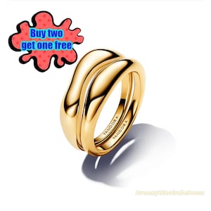 May include: Two gold-colored rings stacked together, with the word "PANDORA" engraved on the side of each ring. The rings have a smooth, curved design. A red and black graphic with the text "Buy two get one free" is in the upper left corner.