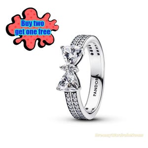 Könnte beinhalten: Ein silberner Ring mit zwei klaren, dreieckigen Edelsteinen und kleineren Akzentsteinen. Das Band hat eine Reihe kleiner Steine. Das Wort "PANDORA" ist auf der Seite des Rings zu sehen. Eine rot-schwarze Grafik mit dem Text "Buy two get one free" befindet sich im Hintergrund.