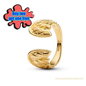 May include: A gold-colored ring with a unique open design. The ring features two stylized leaf-like shapes that curve around the finger. The text "Buy two get one free" is in a speech bubble. The ring is on a white background.