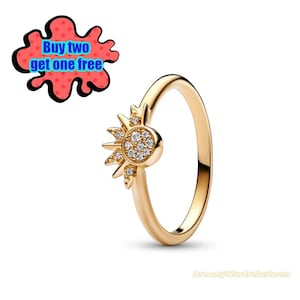 May include: A gold-colored ring with a sunburst design. The sunburst features a central circle of small, clear stones surrounded by pointed rays. The ring is set against a white background. Text in the image reads "Buy two get one free."