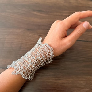 Può includere: Un bracciale a fascia larga realizzato in rete metallica color argento. La struttura presenta un motivo aperto a rete con piccole perline trasparenti e riflettenti integrate nella trama, creando un aspetto strutturato e scintillante.
