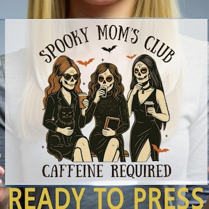 K&ouml;nnte beinhalten: Ein Transferdruck mit drei Figuren mit Totenkopf-Make-up, die Kaffeetassen und ein Buch halten, dazu eine schwarze Katze. Der Text lautet SPOOKY MOM'S CLUB und CAFFEINE REQUIRED, verziert mit kleinen Flederm&auml;usen auf einer transparenten Folie.