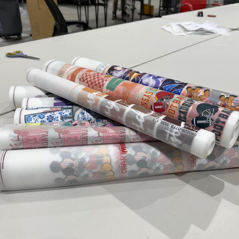 K&ouml;nnte beinhalten: Eine Sammlung von gerollten DTF-Transferfolien mit verschiedenen grafischen Designs, darunter Football-Helme, Zeichentrickfiguren und florale Muster. Die durchscheinenden wei&szlig;en Folienrollen zeigen farbenfrohe Drucke mit Texten wie Kansas City und Logos.
