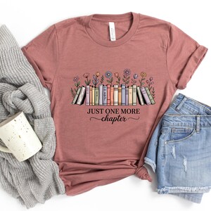 May include: Dusty rose t-shirt with a colourful illustration of books and flowers. The text "JUST ONE MORE chapter" is printed below the books. The shirt is paired with denim shorts and a grey sweater.