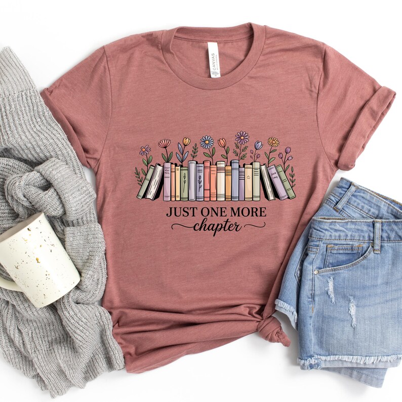 May include: Dusty rose t-shirt with a colourful illustration of books and flowers. The text "JUST ONE MORE chapter" is printed below the books. The shirt is paired with denim shorts and a grey sweater.