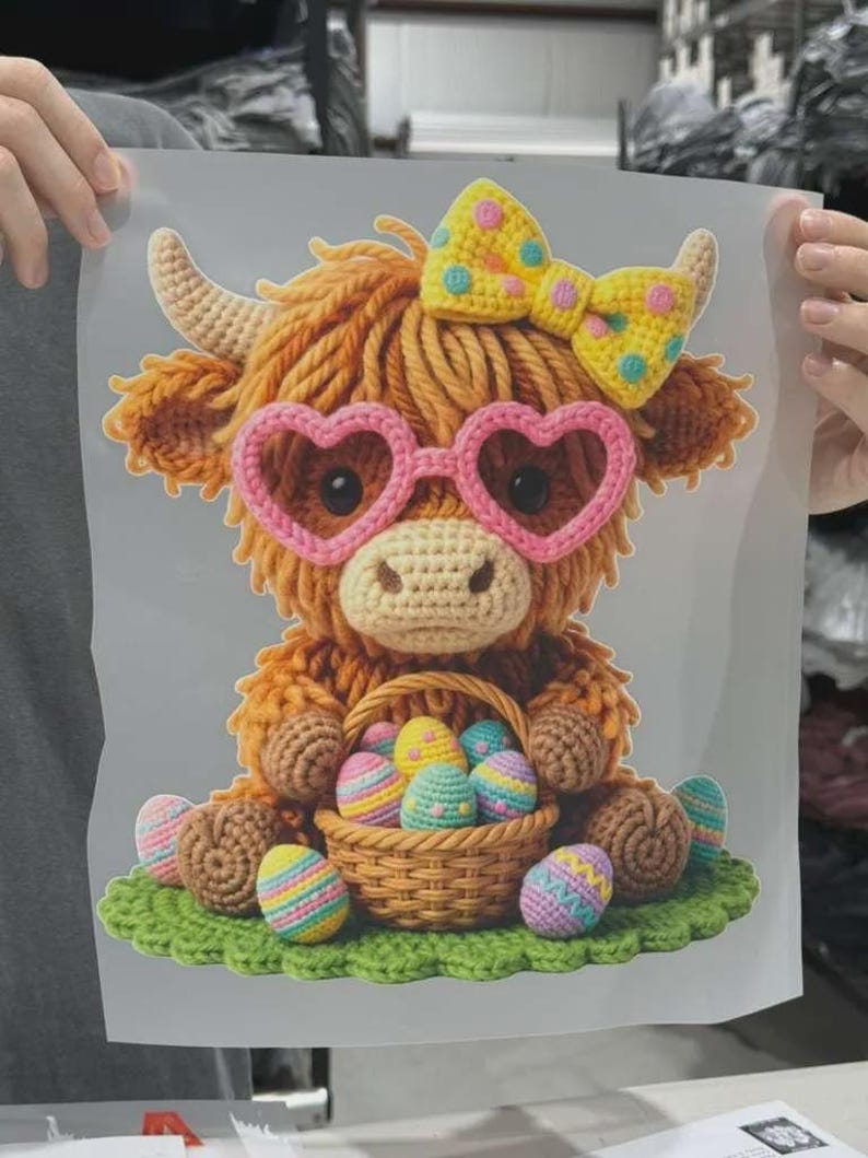 K&ouml;nnte beinhalten: Ein Grafikdruck im H&auml;kel-Stil zeigt ein flauschiges orangefarbenes Hochlandrind mit einer rosa herzf&ouml;rmigen Brille und einer gelben gepunkteten Schleife. Es sitzt auf einer gr&uuml;nen Basis und h&auml;lt einen Korb voller bunter Ostereier.