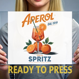 May include: A rectangular print featuring the orange Aperol logo and the text DAL 1919. Below is an illustration of an orange cocktail in a stemmed glass with ice and a straw, surrounded by three oranges with green leaves. Text reads ITALIA SPRITZ.