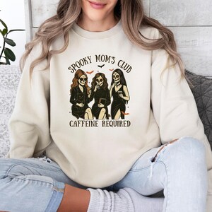 K&ouml;nnte beinhalten: Ein beigefarbenes Sweatshirt mit Rundhalsausschnitt und einem Grafikdruck von drei Figuren mit Totenkopfgesichtern, langem Haar und schwarzen Kleidern. Der Text lautet SPOOKY MOM'S CLUB und CAFFEINE REQUIRED, erg&auml;nzt durch kleine orange Flederm&auml;use.