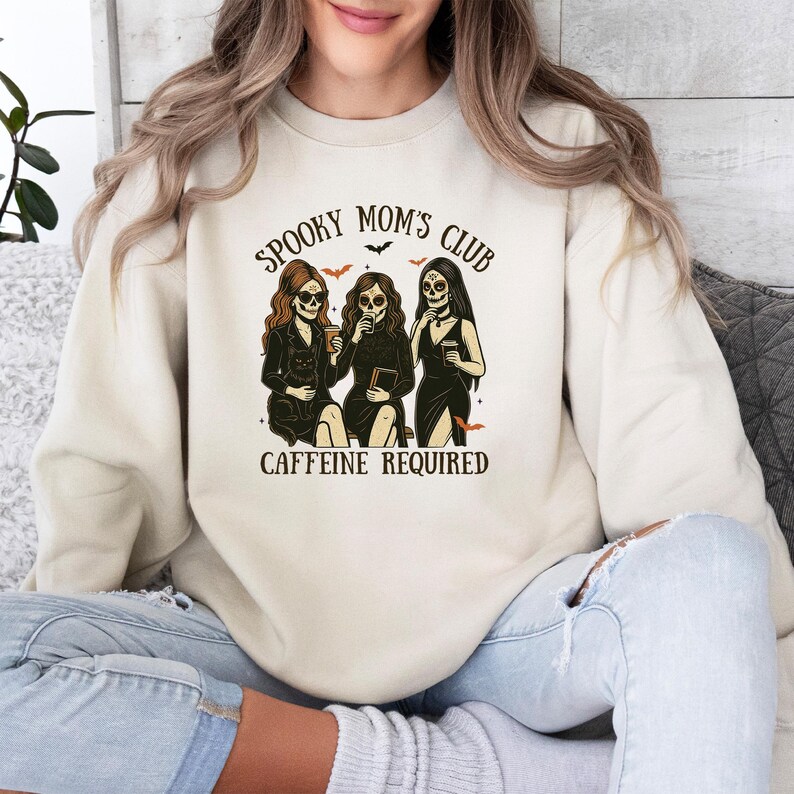 K&ouml;nnte beinhalten: Ein beigefarbenes Sweatshirt mit Rundhalsausschnitt und einem Grafikdruck von drei Figuren mit Totenkopfgesichtern, langem Haar und schwarzen Kleidern. Der Text lautet SPOOKY MOM'S CLUB und CAFFEINE REQUIRED, erg&auml;nzt durch kleine orange Flederm&auml;use.