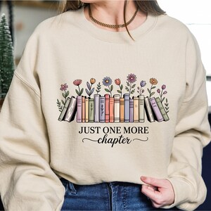 May include: Cream-coloured sweatshirt featuring a graphic of books, flowers, and the text "JUST ONE MORE chapter". The design is printed on the front of the sweatshirt, with a row of books and flowers.