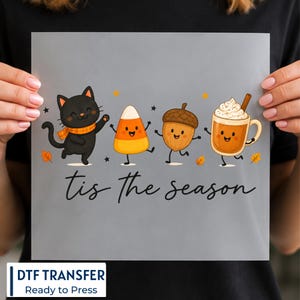 May include: A square DTF transfer sheet featuring a black cat wearing an orange scarf, a piece of candy corn, an acorn, and a mug of coffee with whipped cream and a cinnamon stick. The text 'tis the season' is printed in black cursive below.