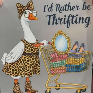 K&ouml;nnte beinhalten: Ein Grafikdruck auf hellgrauem Hintergrund zeigt eine wei&szlig;e Gans in einem Kleid, Kopfschmuck und Stiefeln mit Leopardenmuster sowie einer runden Brille. Die Gans schiebt einen goldenen Einkaufswagen mit einem Spiegel, Schuhen und Kleidung.