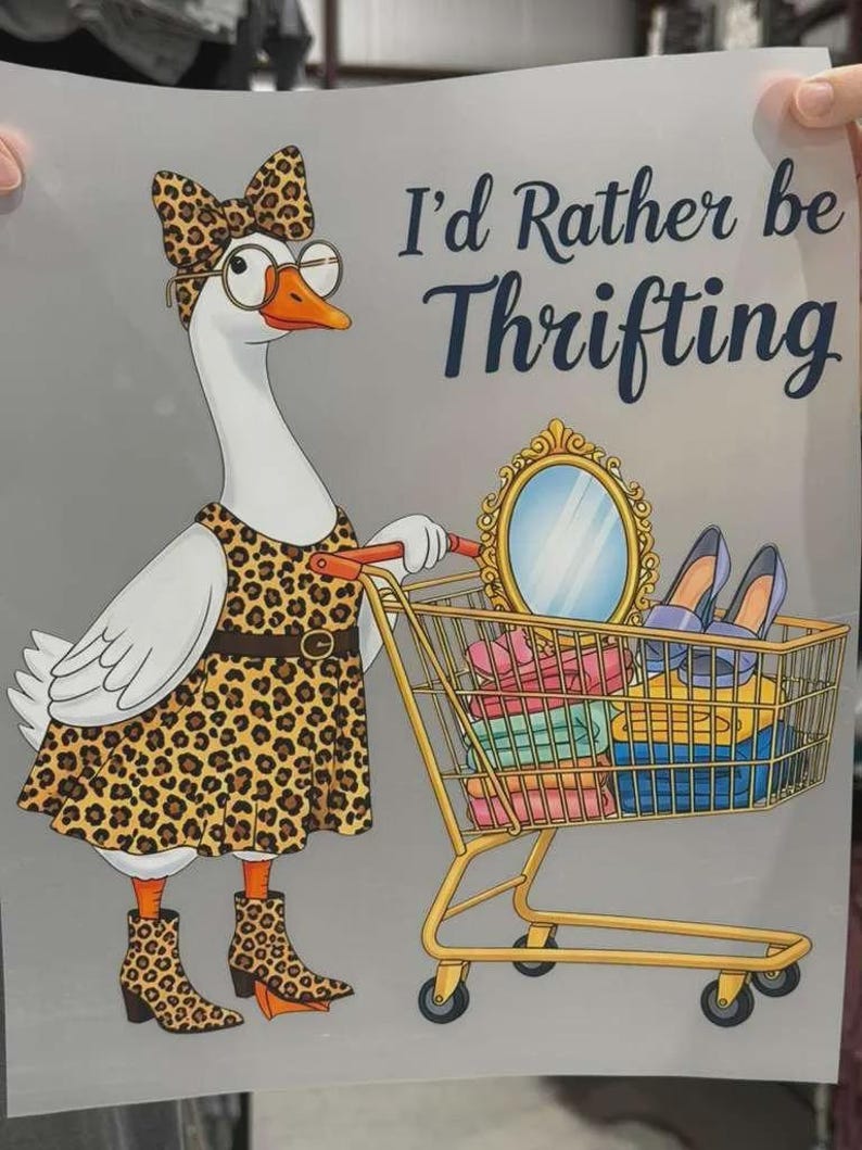 K&ouml;nnte beinhalten: Ein Grafikdruck auf hellgrauem Hintergrund zeigt eine wei&szlig;e Gans in einem Kleid, Kopfschmuck und Stiefeln mit Leopardenmuster sowie einer runden Brille. Die Gans schiebt einen goldenen Einkaufswagen mit einem Spiegel, Schuhen und Kleidung.