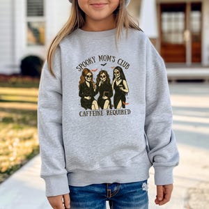 K&ouml;nnte beinhalten: Ein hellgraues Sweatshirt mit Rundhalsausschnitt und einem Grafikdruck von drei Figuren mit Totenkopfgesichtern. Der Text "SPOOKY MOM'S CLUB" steht oben und "CAFFEINE REQUIRED" unten, erg&auml;nzt durch kleine Fledermausillustrationen und Kaffeetassen.