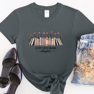 May include: Dark grey t-shirt with a colourful illustration of books and flowers, and the text "Just One More Chapter". The shirt is styled with denim jeans and black heels.
