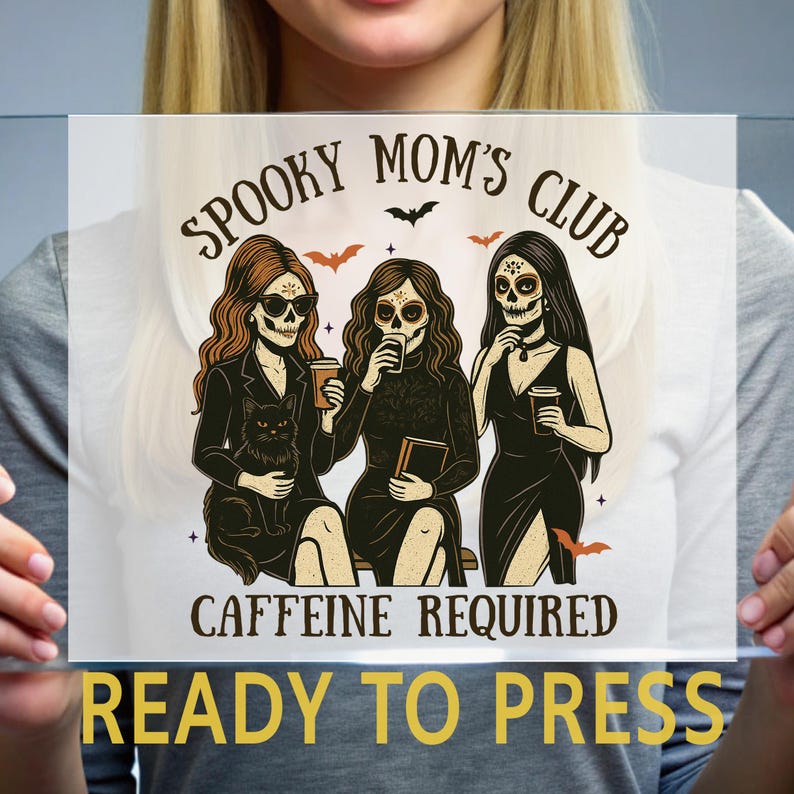 K&ouml;nnte beinhalten: Ein Transferdruck mit drei Figuren mit Totenkopf-Make-up, die Kaffeetassen und ein Buch halten, dazu eine schwarze Katze. Der Text lautet SPOOKY MOM'S CLUB und CAFFEINE REQUIRED, verziert mit kleinen Flederm&auml;usen auf einer transparenten Folie.