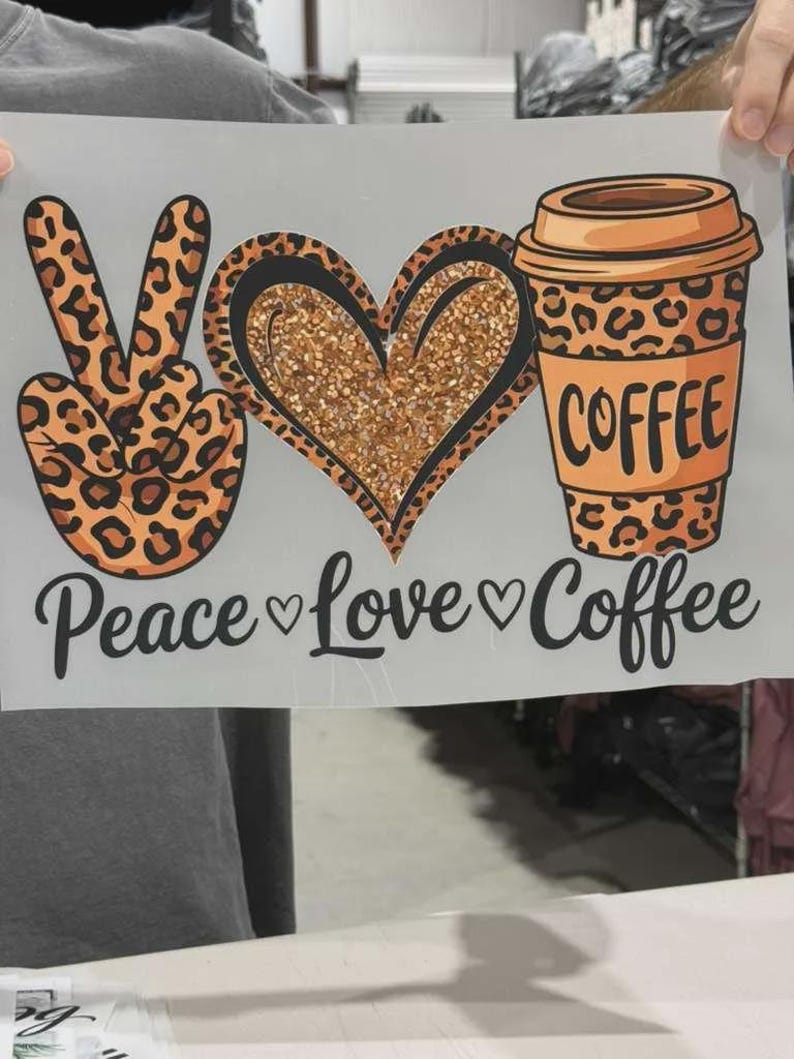 K&ouml;nnte beinhalten: Ein Grafik-Transferbogen mit einem Peace-Zeichen im Leopardenmuster, einem Herz mit goldener Glitzertextur und einem Kaffeebecher im Leopardenmuster. Darunter steht in schwarzer Schrift Peace, Love, and Coffee, getrennt durch kleine Herzen.