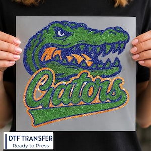 May include: A glittery DTF transfer featuring a stylized green and blue alligator head with orange teeth. Below the alligator, the word Gators is written in a bold, cursive green font with an orange outline, all set against a clear background.