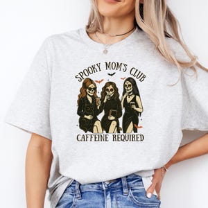 K&ouml;nnte beinhalten: Ein hellgraues T-Shirt mit einer Grafik von drei Figuren mit Totenkopfgesichtern, die schwarze Kleider tragen und Kaffeetassen sowie ein Buch halten. Der Text lautet SPOOKY MOM'S CLUB und CAFFEINE REQUIRED, verziert mit kleinen Flederm&auml;usen und Sternen.