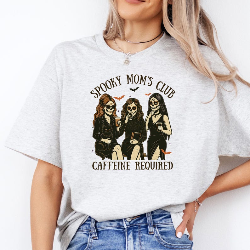 K&ouml;nnte beinhalten: Ein hellgraues T-Shirt mit einer Grafik von drei Figuren mit Totenkopfgesichtern, die schwarze Kleider tragen und Kaffeetassen sowie ein Buch halten. Der Text lautet SPOOKY MOM'S CLUB und CAFFEINE REQUIRED, verziert mit kleinen Flederm&auml;usen und Sternen.