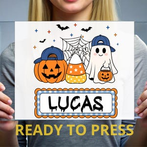 May include: A white rectangular graphic print featuring a jack-o'-lantern wearing a blue baseball cap, a candy corn, and a ghost wearing a blue baseball cap holding a small pumpkin bucket. Below, a blue checkered frame contains the name LUCAS.