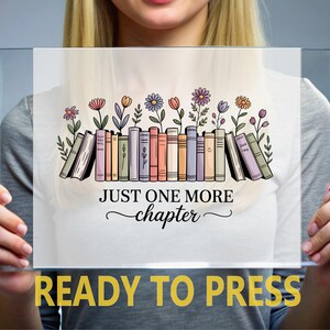 May include: A clear rectangular panel with the text "JUST ONE MORE chapter" and "READY TO PRESS". The design features a row of colourful books with flowers and foliage above them. The panel is held by a person.