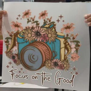 K&ouml;nnte beinhalten: Ein W&auml;rme&uuml;bertragungsaufkleber mit einer t&uuml;rkisfarbenen Kamera, umgeben von rosa und pfirsichfarbenen Blumen, kleinen Sternakzenten und dem Text "Focus on the Good" in brauner Schreibschrift unten auf einem wei&szlig;en Blatt.