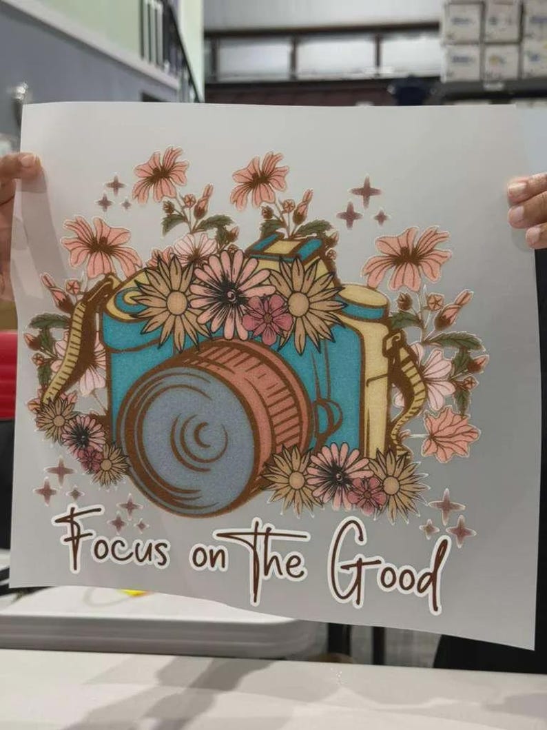 K&ouml;nnte beinhalten: Ein W&auml;rme&uuml;bertragungsaufkleber mit einer t&uuml;rkisfarbenen Kamera, umgeben von rosa und pfirsichfarbenen Blumen, kleinen Sternakzenten und dem Text "Focus on the Good" in brauner Schreibschrift unten auf einem wei&szlig;en Blatt.