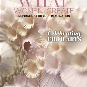 以下が含まれることがあります： ピンク色の文字で「WHAT WOMEN CREATE」と書かれた雑誌の表紙。表紙には、白とクリーム色の繊維芸術のクローズアップがあり、右下隅に「Celebrating FIBER ARTS」という言葉があります。