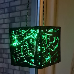 Könnte beinhalten: Eine würfelförmige Lampe, die an Ketten aufgehängt ist und ein kompliziertes, grün beleuchtetes Design aufweist. Die Lampe befindet sich vor einem Fenster. Das komplexe Muster leuchtet hell und erzeugt einen auffälligen visuellen Effekt.