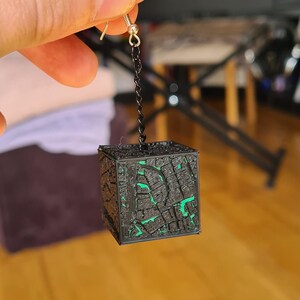 May include: A black cube-shaped earring with intricate carved details and green accents. The earring hangs from a silver hook and chain. The cube design is reminiscent of a puzzle box.