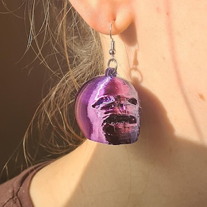 May include: A close-up of a purple and black skull-shaped earring. The earring is suspended from a silver hook. The earring is worn by a person. The person is wearing a brown shirt.