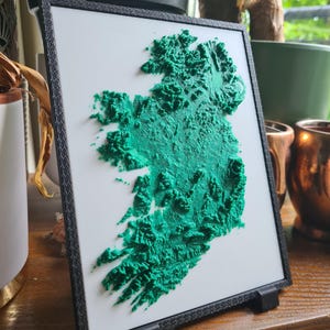May include: A framed 3D map of Ireland, showing the island's topography in green against a white backdrop. The map is set within a black frame and stands on a wooden surface. The relief details the island's topography.