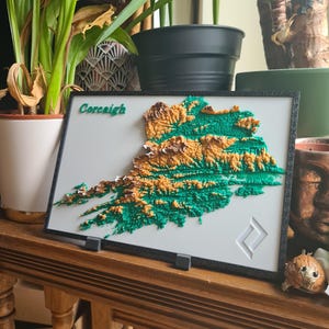 May include: A framed 3D topographical map of an area, labelled "Corcaigh" in green. The map shows green and brown elevations on a white background, within a black frame. The map is displayed on a wooden surface, with a decorative symbol in the corner.