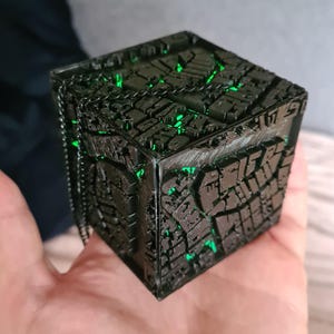 May include: A black, cube-shaped object with intricate, geometric patterns and glowing green accents. The object has a chain attached to it. The design suggests a futuristic or sci-fi theme.