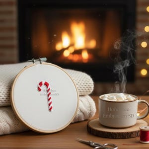 Può includere: Un telaio da ricamo con un motivo a bastoncino di zucchero a righe rosse e bianche. Una tazza di cioccolata calda con marshmallow è su un sottobicchiere di legno. Il testo "Honeybee EmbroiderySD" è visibile.