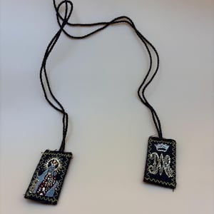 May include: A black cord with two rectangular fabric charms. One charm features an embroidered image of a woman in a crown, the other charm features an embroidered crown and the letters "M.R."