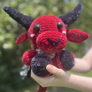 May include: A handmade red crochet bull plushie with black horns, hooves, and accents. The plushie has black button eyes and a white and gray tail. The bull is held in a person's hands against a blurred green background.