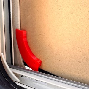 Op de afbeelding: Close-up van een rode plastic raamvergrendeling. De gebogen vergrendeling bevindt zich in de buurt van de rand van een raamkozijn. Het frame is zwart en wit, met een lichtbruin paneel op de achtergrond.