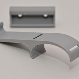 Könnte beinhalten: Eine matte graue Kunststoffhalterung bestehend aus einem geschwungenen Arm mit einem rechteckigen Schlitz und einem separaten rechteckigen Montageteil mit zwei runden Schraublöchern. Das Design umfasst eine zentrale Stützstruktur und ein Hakenende.