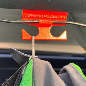 Può includere: Un gancio arancione brillante stampato in 3D, progettato per appendere oggetti, è sospeso a un filo nero. Il gancio ha due sezioni inferiori curve e una sezione superiore con il testo "3DvoxelWorks.sa". Una giacca è appesa al gancio.