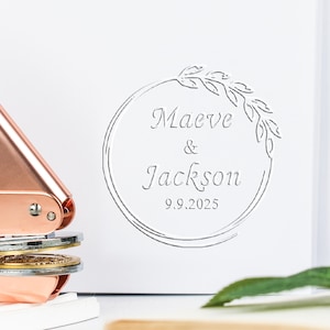 May include: A rose gold embosser next to a white notebook with the names "Maeve & Jackson" and the date "9.9.2025" in a decorative circle. The notebook has a spiral binding.