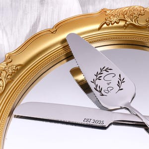 Puede incluir: Juego de servidor y cuchillo para pastel plateado con detalles grabados. El servidor presenta un diseño de hojas decorativas con las iniciales "C & G". El cuchillo está grabado con "EST.2025". El juego se muestra en un espejo dorado.