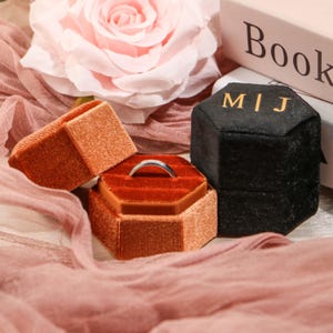 May include: A silver ring sits in an open, hexagonal, velvet ring box. The box is a deep orange colour, with a contrasting red interior. A second orange box and a black box with gold lettering are also visible.