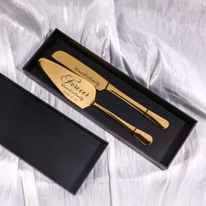 Könnte beinhalten: Ein goldfarbenes Tortenheber- und Messerset in einer schwarzen Box. Der Tortenheber ist mit dem Wort "Forever" und dem Datum "2025-5-21" graviert. Messer und Tortenheber sind mit den Namen "David & Emily" graviert.