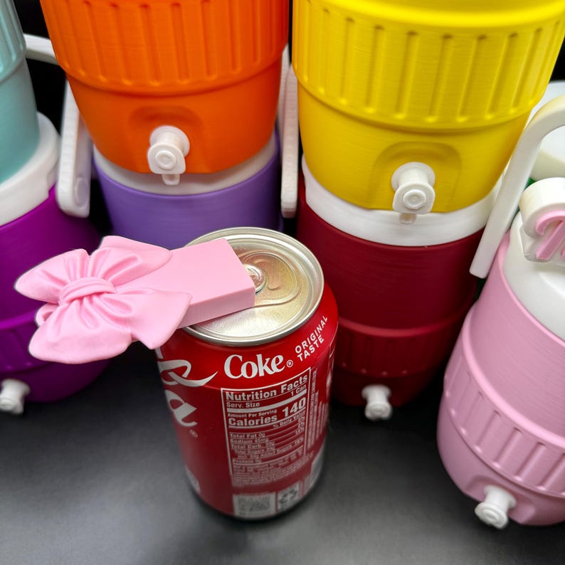 May include: A red Coca-Cola can with a pink bow and a pink rectangular object on top. Several colorful water coolers are stacked in the background, including orange, yellow, purple, and pink coolers.