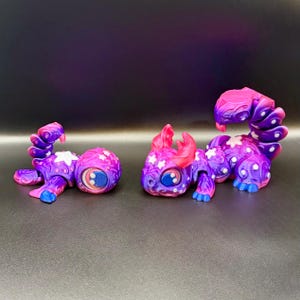 May include: Two whimsical, purple and pink fantasy creatures with star and dot patterns. One is lying down, the other is upright. Both have large blue eyes, pink horns, and blue feet.