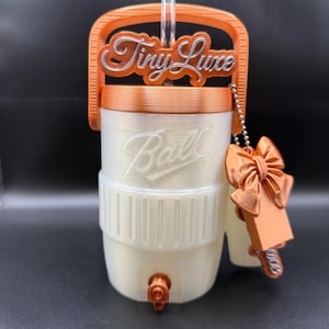 Puede incluir: Un dispensador de bebidas cilíndrico blanco con el logotipo Ball en relieve en la parte frontal, con asa, tapa y grifo de color naranja. El asa superior muestra el texto Tiny Luxe en plata. Un dije de lazo naranja cuelga de una cadena.