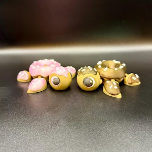 May include: A selection of quirky, 3D-printed eye sculptures in the shape of donuts. The donuts have pink and brown icing with white sprinkles and chocolate drizzle. The eye pupils are detailed with black and white accents.